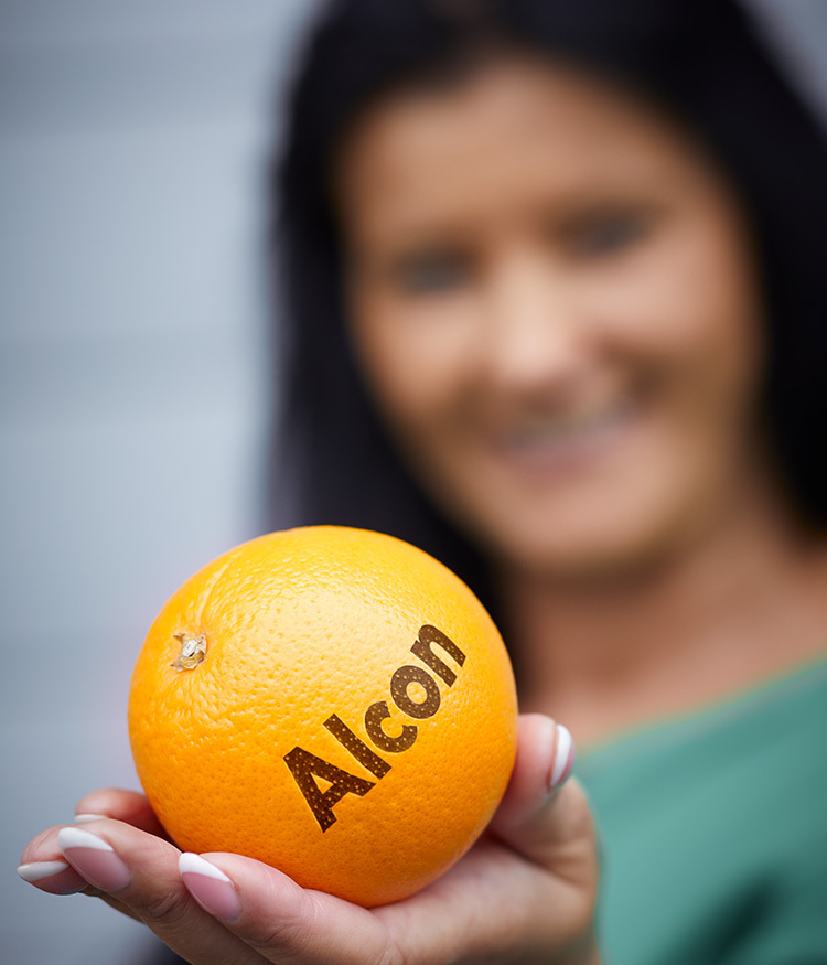 Frau hält eine Orange mit dem Aufdruck „Alcon“ auf der Schale, Fokus auf die Frucht im Vordergrund.