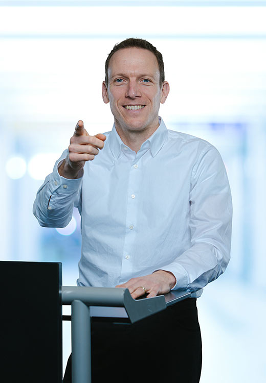 Mann im weißen Hemd, der lächelt und auf die Kamera zeigt, während er hinter einem Rednerpult steht.