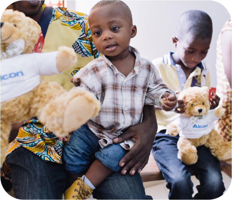 Маленький ребёнок, одетый в клетчатую рубашку и закатанные джинсы, сидит на коленях у взрослого и тянется к плюшевому мишке, одетом в рубашку с логотипом Alcon. На заднем плане сидит ещё один ребёнок, тоже держащий такого же плюшевого мишку, и смотрит на него.