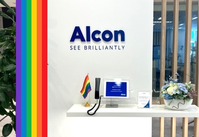 「See Brilliantly」のスローガン付きAlconのロゴがある受付カウンター。タブレット端末と花のアレンジメントの横に小さなレインボープライドフラッグが飾られている。画像の左端には虹色の縁取りがある。