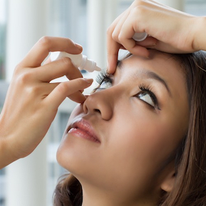 Młoda kobieta o brązowych włosach odchyla głowę do tyłu i zakrapla krople do lewego oka.