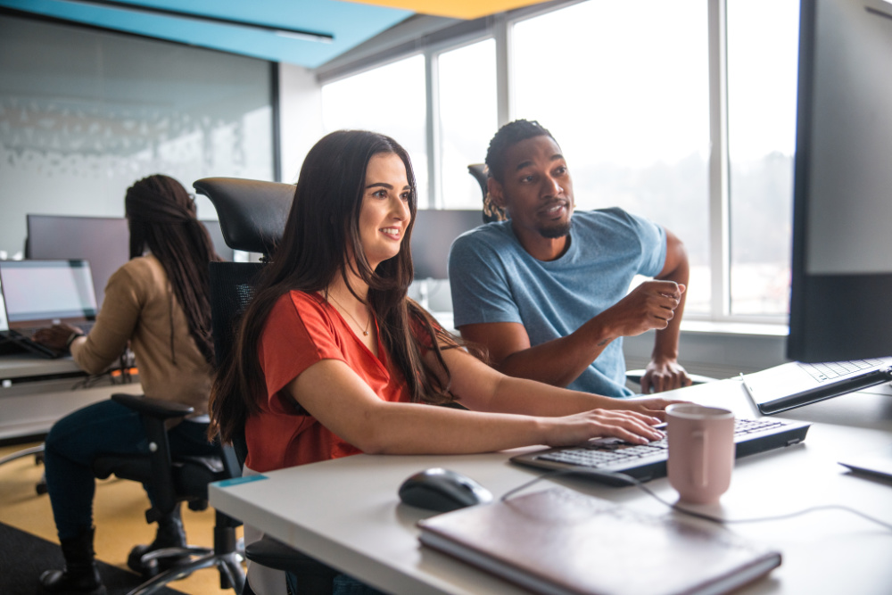 A woman is typing on a computer while a man looks on and gestures with his hands. They are working at a desk in a modern office.