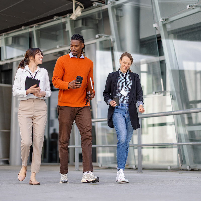 Three people are walking forward, appearing to head towards a modern building. One person holds a tablet, another holds a coffee cup, and the third wears a blazer and holds a badge.
