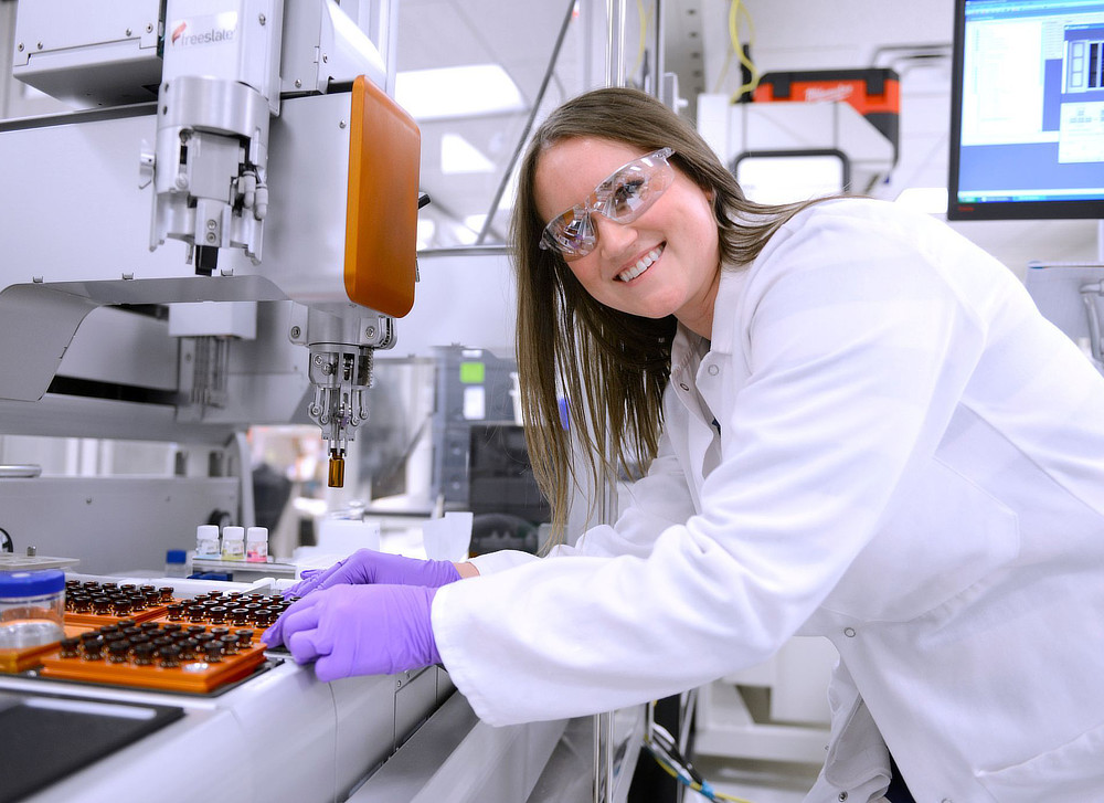 A female scientist in a lab coat and safety glasses smiles while working with automated laboratory equipment. She's wearing purple gloves and operating a machine with a robotic arm, likely involved in sample analysis or research.
