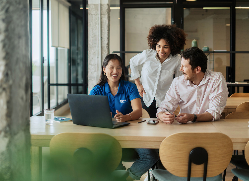 Three professionals, including a woman in an Alcon polo shirt, collaborate around a table in a bright office.