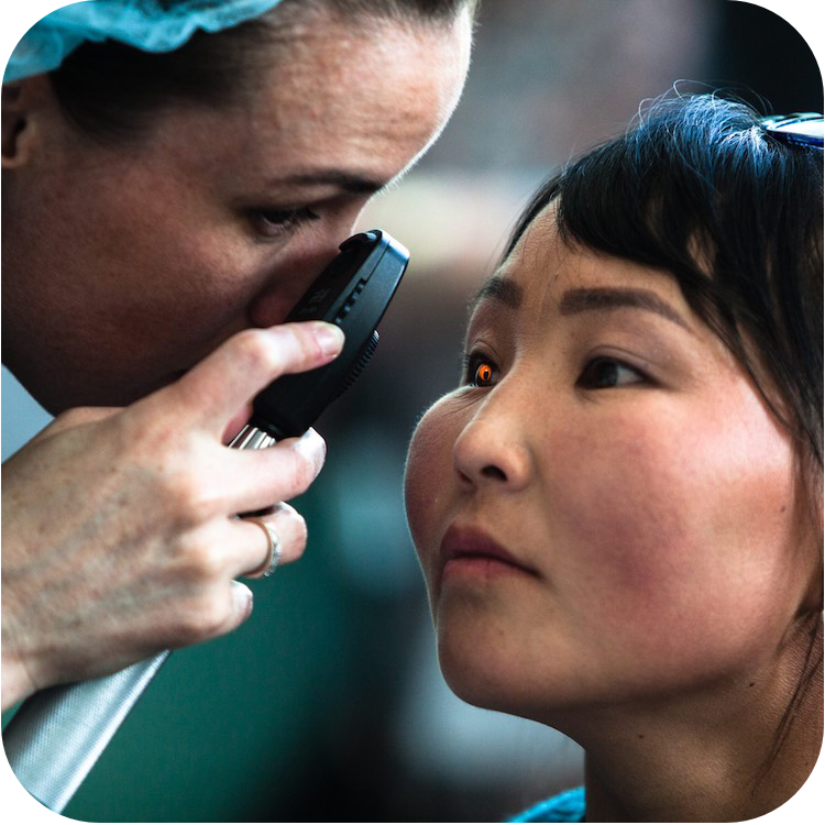 Офтальмолог осматривает глаз пациента с помощью ручного офтальмоскопа. Пациентка смотрит прямо перед собой, а специалист наклоняется для детального осмотра. Офтальмоскоп испускает слабый оранжевый свет, отражающийся в глазу пациента.
