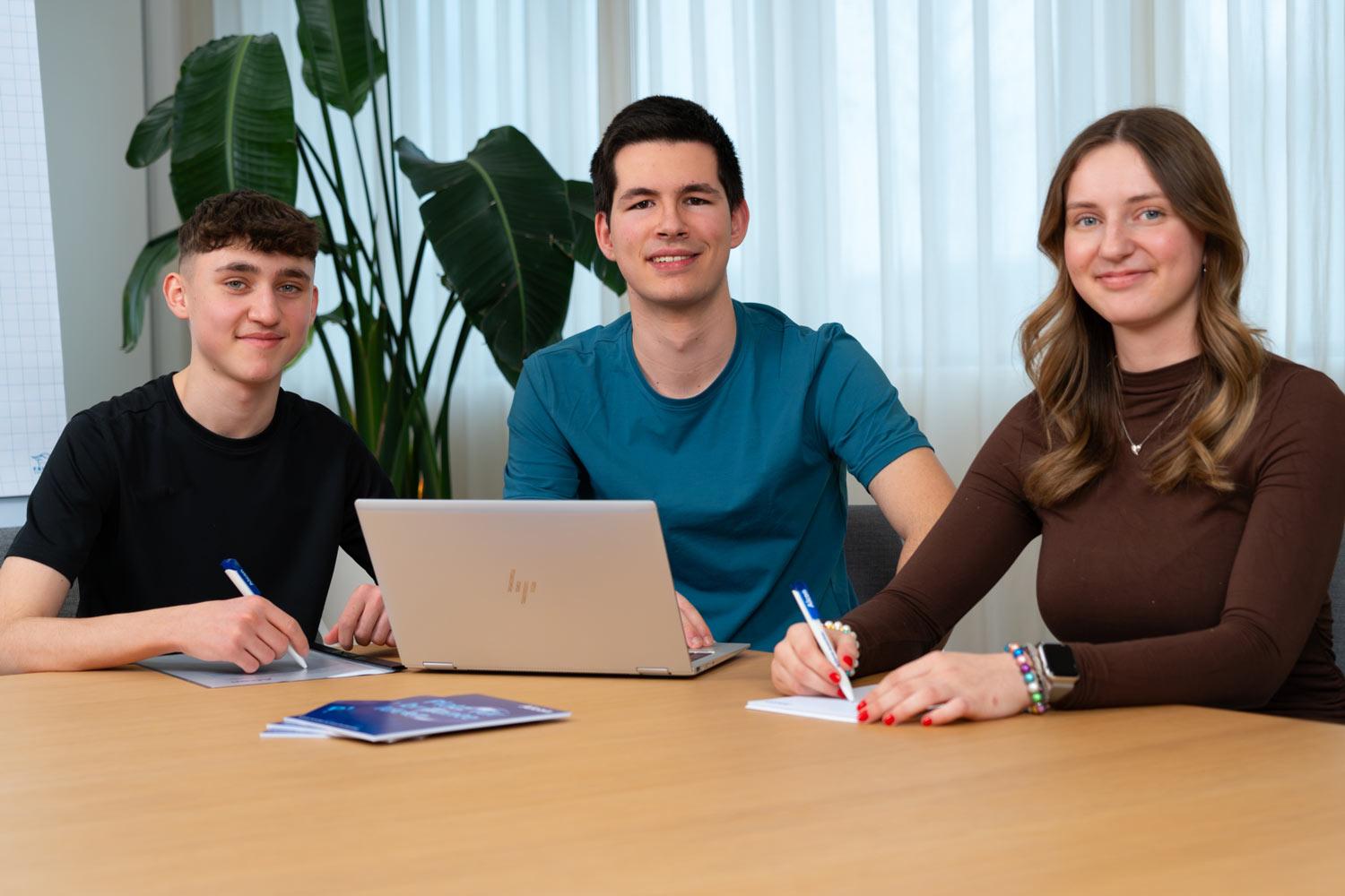 Drei junge Fachkräfte sitzen gemeinsam an einem Tisch mit Laptop und Notizblöcken und arbeiten in einer hellen Büroumgebung zusammen.