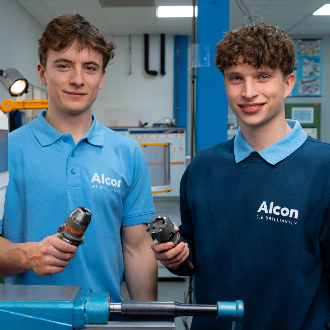 Zwei junge Männer in Alcon-Uniformen halten Werkzeuge in einer Werkstatt und lächeln in die Kamera.