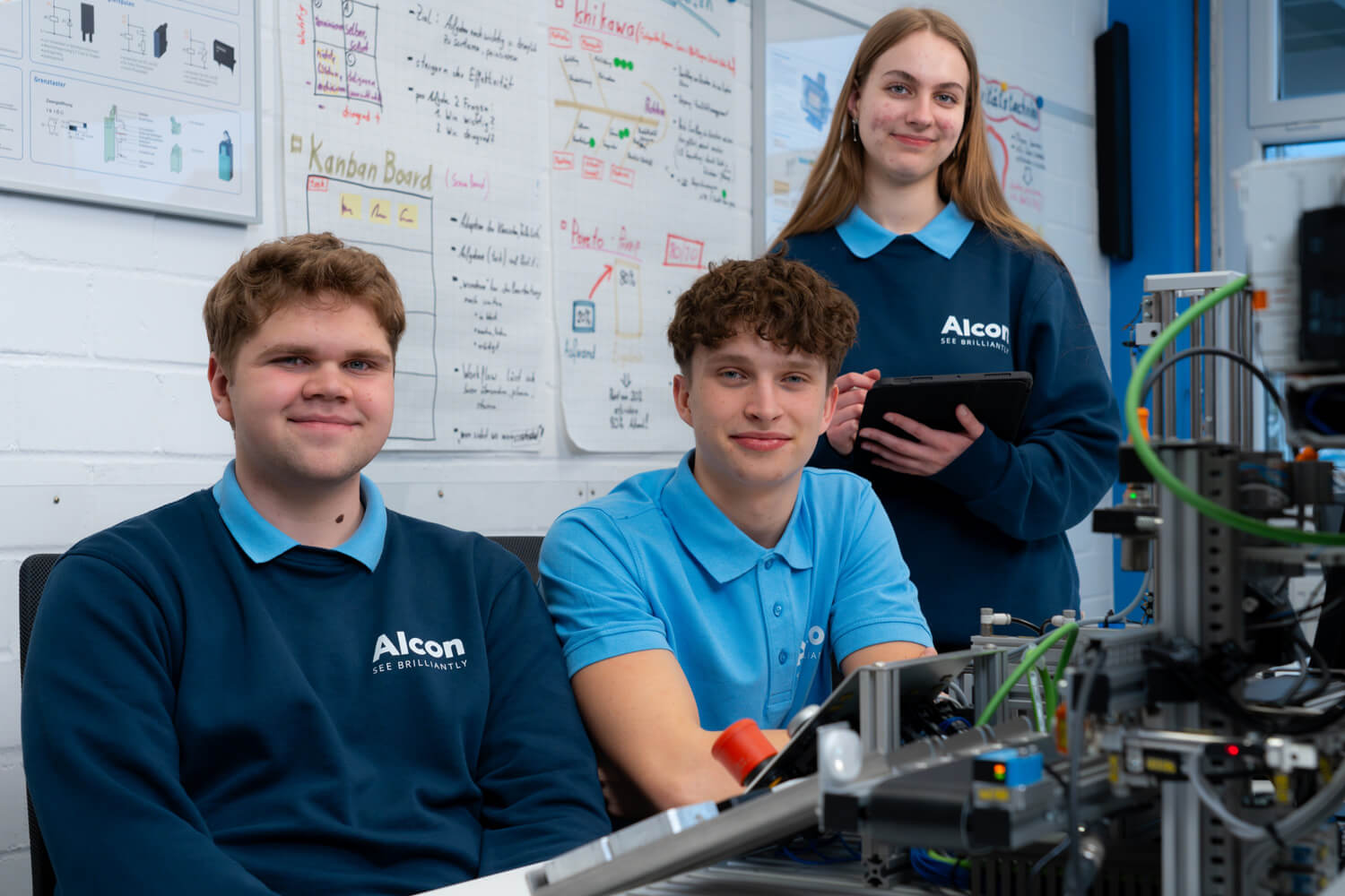 Drei junge Alcon-Auszubildende arbeiten gemeinsam in einer technischen Ausbildungsumgebung, einer hält ein Tablet, die anderen sitzen an Geräten vor Projektplakaten.