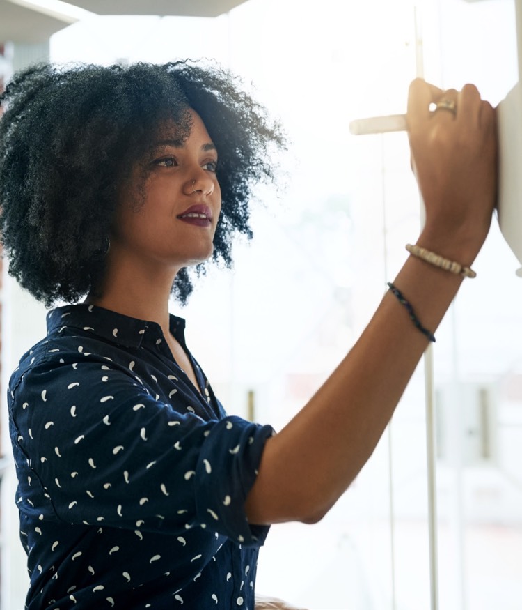 Kıvırcık saçlı ve desenli lacivert bluz giyen bir kadın, iyi aydınlatılmış bir alanda beyaz tahtaya yazıyor veya çizim yapıyor.