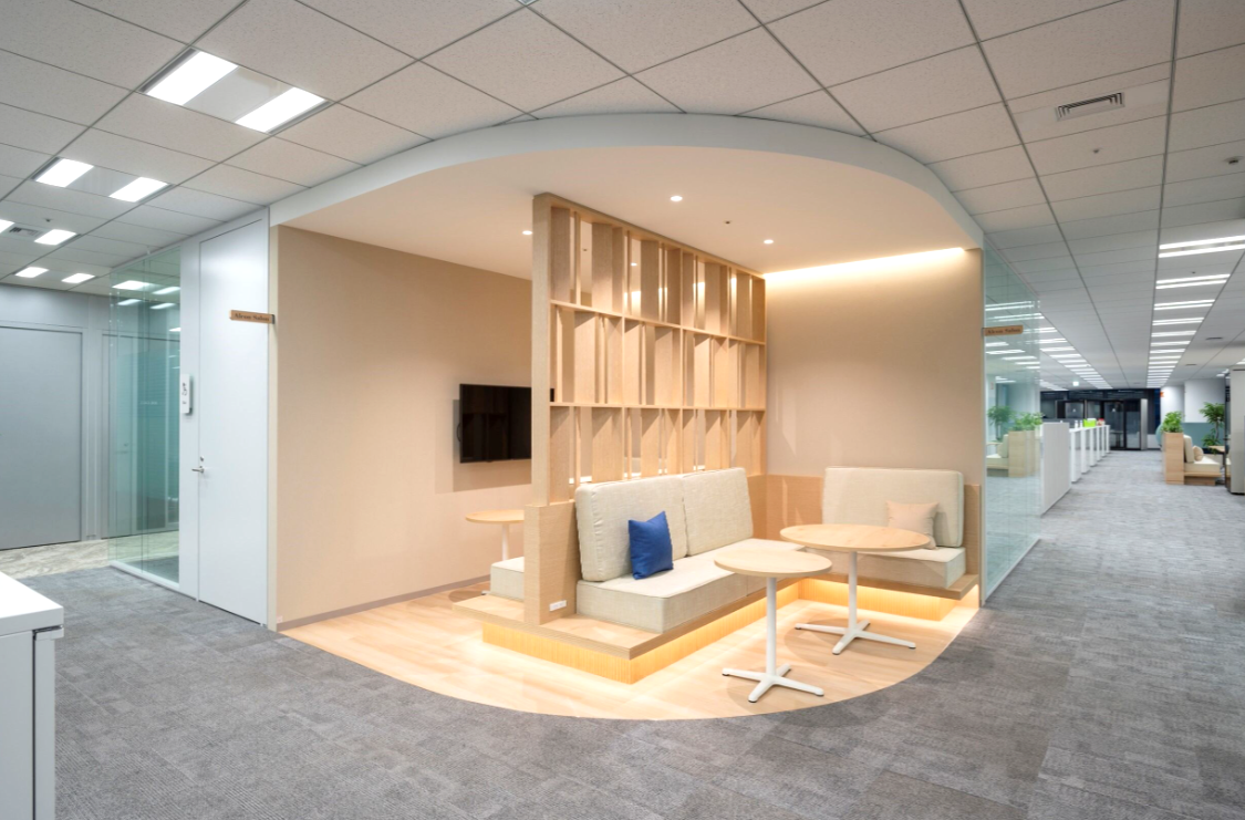 明るい木材を基調としたモダンなオフィス内の半個室型の休憩スペース。クッション付きベンチと丸テーブルが設置され、柔らかな照明が照らしています。