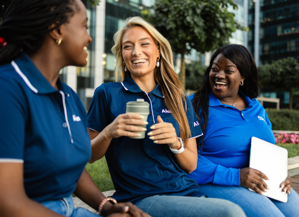 Three women, all wearing blue Alcon-branded polo shirts, are seated outdoors and laughing. One holds a drink, and another holds a tablet.