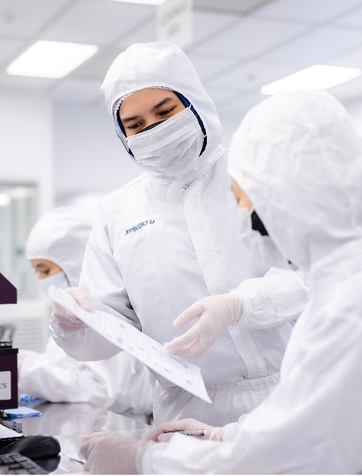 A scientist wearing a full white protective suit, mask, and hood works in a sterile cleanroom environment. They are focused on a task with their hands out of view, surrounded by white protective coverings. Another scientist is visible in the background.