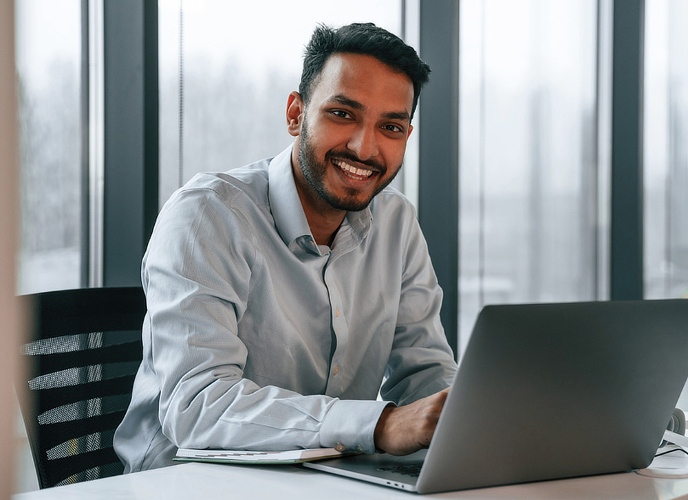 A smiling man with dark hair is sitting at a desk and looking at the camera. He is wearing a light blue dress shirt and working on a laptop.