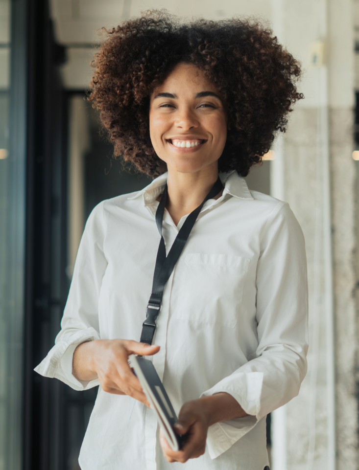 A woman with a long dark hair is smiling at the camera while holding a tablet computer. She is wearing a white shirt and a lanyard.