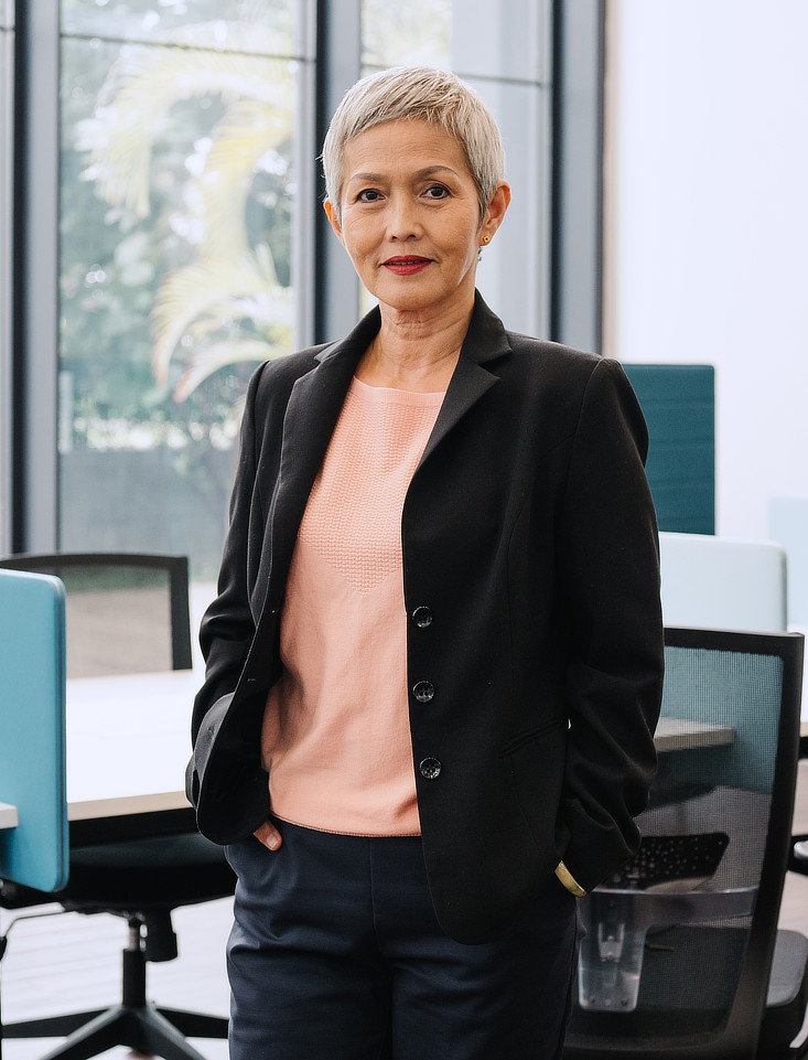 A woman with short, silver hair is standing in what appears to be an office space. She is wearing a black blazer over a light pink shirt and dark jeans.