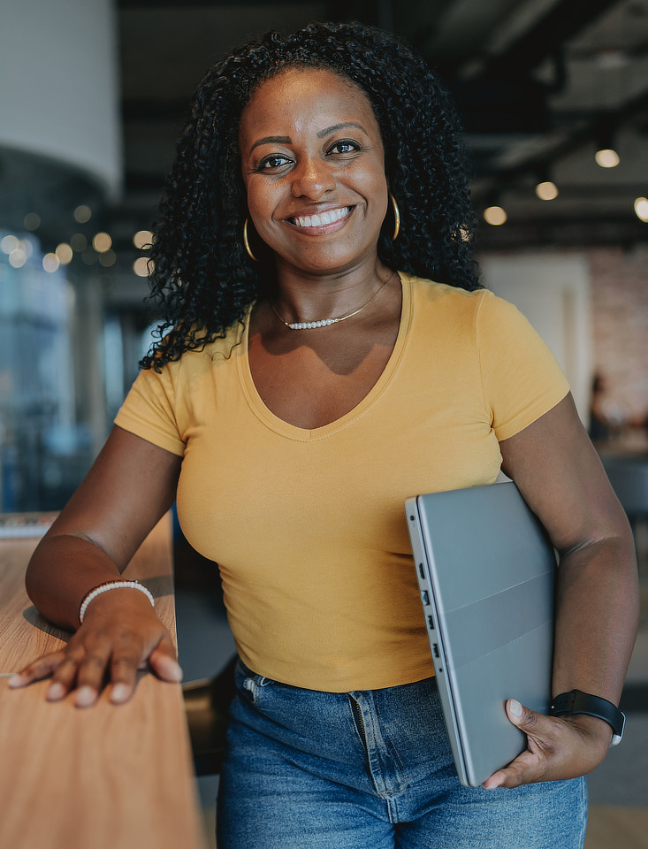 A woman with dark, curly hair is smiling at the camera while leaning on a wooden surface and holding a laptop under her arm. She is wearing a yellow t-shirt and blue jeans.