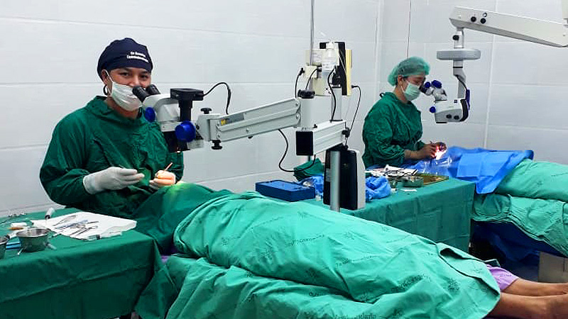 2人の眼科医が医療施設内で手術を行っている様子。手術用顕微鏡を使用し、緑の手術着、マスク、手袋を着用。患者は緑色のシーツをかけられた手術台に横たわっている。