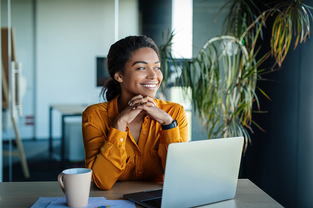 A young Black woman working on a laptop, smiling and looking to the side.