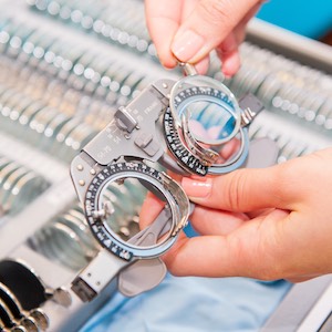 Dłonie trzymające oprawkę próbną do badania wzroku, z okrągłymi, regulowanymi soczewkami. W tle widoczna tacka z różnymi soczewkami.