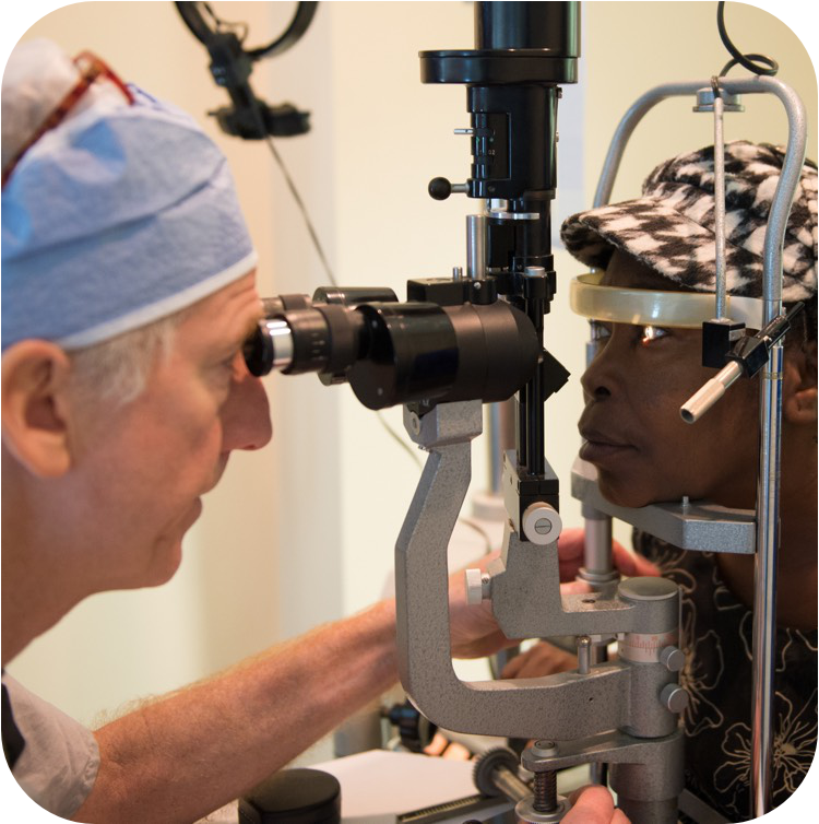 Врач в голубой хирургической шапочке проводит осмотр глаз с помощью щелевой лампы-биомикроскопа. Пациентка опирается подбородком на опору прибора и смотрит прямо перед собой.