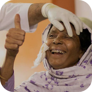 Радостная пожилая женщина в цветочном платке улыбается, пока медицинский работник в перчатках осторожно осматривает её глаз. Она поднимает большой палец вверх, выражая радость и благодарность. Под платком заткнута салфетка, что указывает на недавнюю медицинскую процедуру.