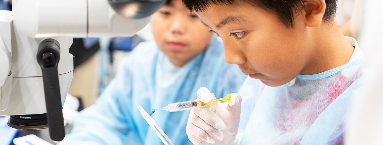 青いガウンと手袋を着用した少年が、サイエンスまたは医療体験ワークショップでシリンジを慎重に扱っている様子。背景には同じような服装の別の子どもが見守っている。