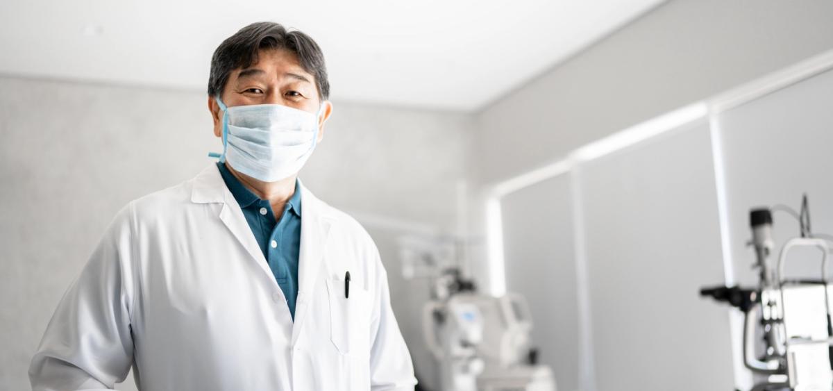 Eye care professional in lab coat and mask, standing in examination room