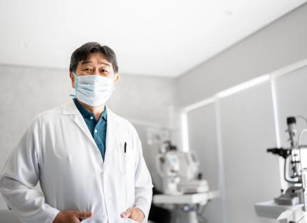 Eye care professional in lab coat and mask, standing in examination room