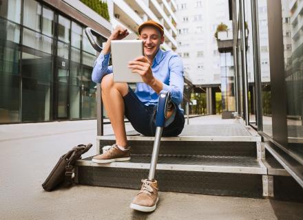 Young man with a prosthetic leg, sitting on stairs outside office building, smiling and looking at tablet