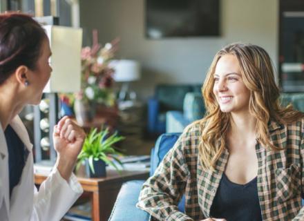 A healthcare professional in a white coat engages in conversation with a smiling woman in a plaid shirt, sitting in a cozy, well-lit waiting area with green plants and comfortable furniture.
