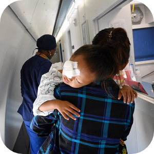 Маленький ребёнок с повязкой на глазу кладёт голову на плечо женщины пока его несут по медицинскому учреждению. Женщина в клетчатой рубашке идёт по узкому коридору, а на заднем плане виден медицинский работник в хирургическом халате. Сцена говорит о послеоперационном уходе или восстановлении.