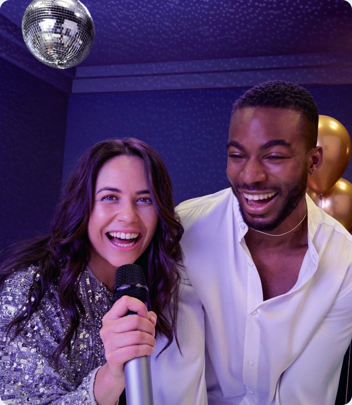Woman holding a microphone and smiling next to a man, both laughing under a disco ball at a party with balloons in the background