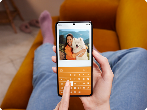 Person holding a smartphone while sitting on a couch, viewing a photo of a woman hugging a fluffy white dog, with a calendar interface below the image