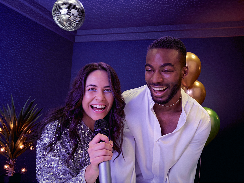 Woman holding a microphone and smiling next to a man, both laughing under a disco ball at a party with balloons in the background
