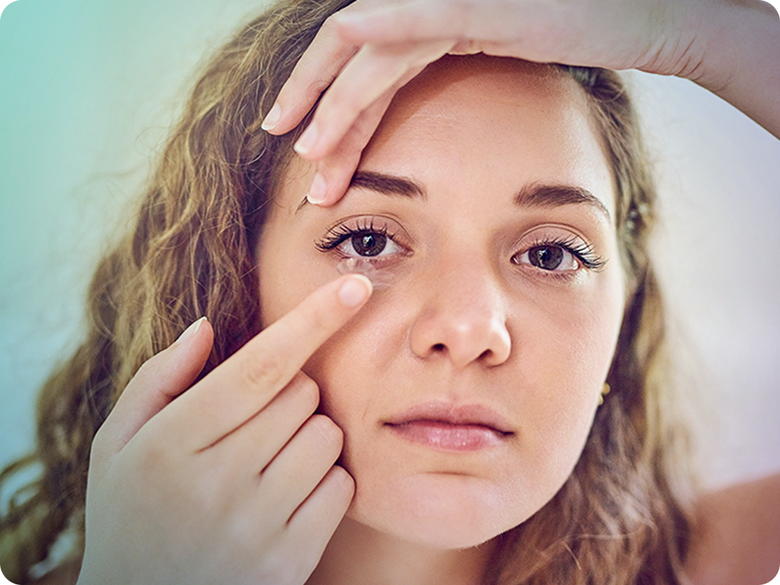 Young woman with wavy blonde hair inserting a contact lens into her eye while holding her upper eyelid open with one hand