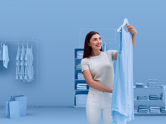 Smiling woman holding up a light blue dress in a modern, monochromatic blue-toned boutique with neatly organized clothing racks and shelves in the background