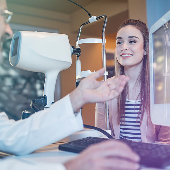 Young woman smiling during an eye exam, sitting across from an eye care professional using diagnostic equipment in a clinic setting